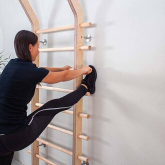 Femme s'étirant en utilisant une barre de traction en bois. Elle est vêtue d'un t-shirt noir et de pantalons de sport noirs, avec une jambe étendue vers le haut, pendant qu'elle tient son pied. En arrière-plan, le mur est clair et il y a une plante à côté