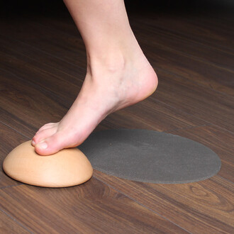 Un pied nu repose sur un accessoire en bois en forme de demi-sphère, placé sur un sol en bois. À côté, on voit un disque de surface grise.