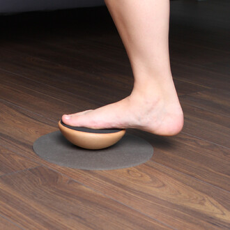 Un pied se repose sur un objet en bois en forme de demi-sphère, qui est sur une surface ronde en matériau gris. L'environnement est intérieur avec un sol en bois.