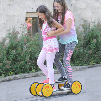 Deux filles s'amusent sur un appareil de jeu Pedalo® Famille. L'une est debout sur la plateforme, tandis que l'autre la maintient par la taille pour l'équilibre. Elles sont entourées de verdure et semblent profiter d'une journée en plein air. Les vêtement