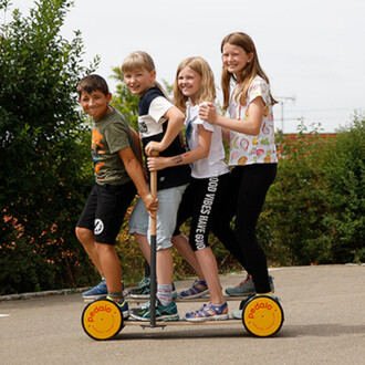 Quatre enfants s'amusent sur un Pedalo® Famille, un véhicule à roulettes. Ils sont positionnés côte à côte, tenant une barre centrale pour se stabiliser. Le décor est extérieur, avec des arbres en arrière-plan. Les enfants sourient et semblent ravis de le