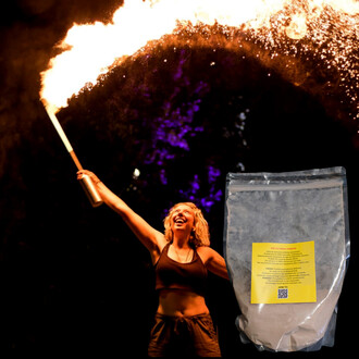 Poudre Liko de 1 litre présentée avec un sachet transparent au premier plan, tandis qu'en arrière-plan, une artiste portant un haut sans manches sourit en exécutant un numéro de jonglage de feu, l'instrument produisant des flammes vives dans la nuit.