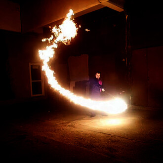 Un artiste jongleur manipule un bâton enflammé, créant une traînée de flammes lumineuses dans un environnement sombre. La scène met en évidence les mouvements dynamiques du jongleur, avec des couleurs vives de feu contrastant avec l'obscurité autour.