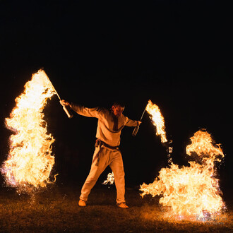 Un artiste de jonglerie utilise des bâtons enflammés, créant de grandes flammes dans l'obscurité. Le spectacle se déroule à l'extérieur, sur une pelouse, alors qu'il exécute des mouvements dynamiques, entouré de feu.