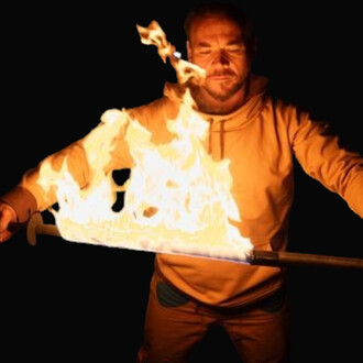 Homme en tenue orange manipulant un bâton enflammé lors d’un spectacle de feu sur fond noir.