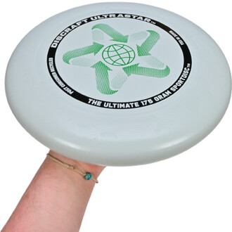 Frisbee Ultrastar phosphorescent blanc tenu dans une main, avec un design en vert et noir, comprenant un motif en forme d'étoile et un globe au centre, avec l'inscription DISCRAFT ULTRASTAR et THE ULTIMATE 175 GRAM SPORTDISC.
