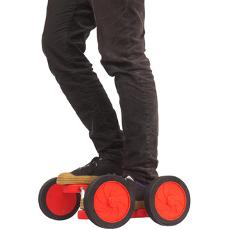 Personne debout sur un Fun Racer Status, planche rouge à quatre roues orange et pneus noirs, portant pantalon noir et baskets noires.