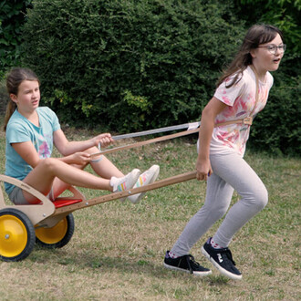 Deux jeunes filles jouent dans un jardin. L'une d'elles est assise dans une charrette en bois, tandis que l'autre la tire avec des sangles. La fille assise porte un t-shirt bleu et shorts, et la fille debout est vêtue d'un t-shirt coloré et d'un pantalon 