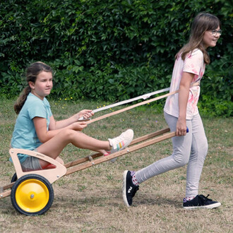 Deux enfants jouent dans un jardin. L'un est assis dans une charrette en bois à roues jaunes, tandis que l'autre tire la charrette avec des cordes. L'enfant assis porte un t-shirt bleu, et l'enfant qui tire porte un t-shirt aux teintes pastel. En arrière-