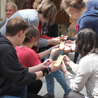 Groupe de jeunes participants tenant des objets semblables à des rampes en bois, en train de s'entraider pour faire rouler une balle ou un objet au milieu d'eux, dans un cadre extérieur, ensoleillé.