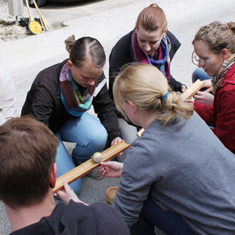 Groupe de personnes accroupies, concentrées sur une activité de jonglerie, avec une barre en bois et un objet sphérique. Certains portent des écharpes colorées. Le cadre est extérieur avec un sol en gravier.