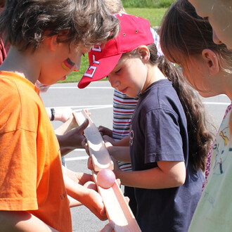 Groupe d'enfants participant à une activité de jonglerie, concentrés sur un parcours en bois, avec une balle rose. Un enfant porte un tee-shirt orange et un autre une casquette rouge. L'ambiance est enjouée et ensoleillée.