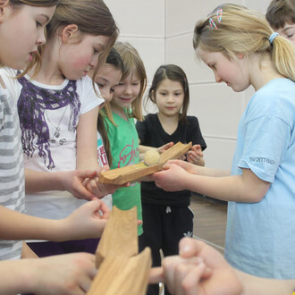 Groupe d'enfants interagissant avec un objet en bois sur lequel repose une balle, montrant leur concentration et collaboration. Ils se tiennent en cercle, certains touchant l'objet et d'autres observant attentivement.