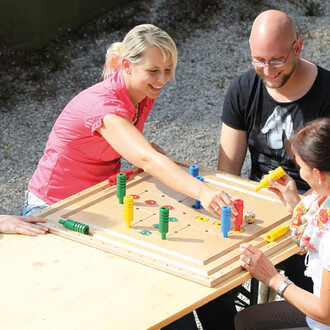 Trois personnes jouent autour d'une table à un jeu de société. Le jeu est en bois, avec des pièces colorées (rouges, jaunes, vertes, et bleues) disposées sur un plateau. L'une des personnes, une femme aux cheveux blonds, sourit en déplaçant une pièce. Un 