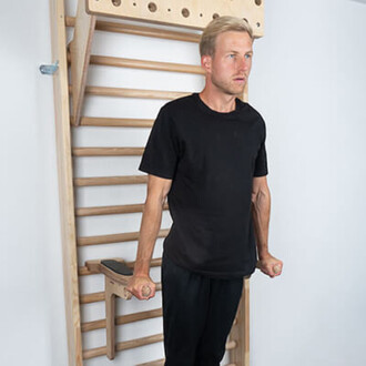 Homme en t-shirt noir effectuant un exercice de musculation sur des barres parallèles, installé devant un mur d'escalade en bois.