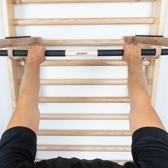 Deux mains tenant une barre de traction fixée à une échelle en bois, sur un mur blanc. Les bras sont visibles, portant un t-shirt noir. La barre est en bois avec des sections noires pour une prise confortable.