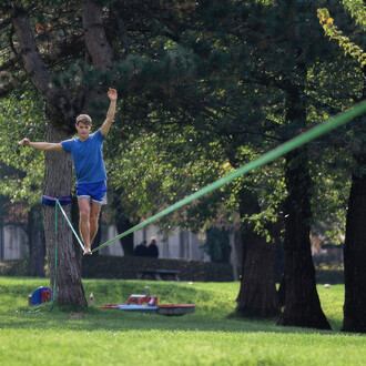 Personne marchant sur une slackline verte du kit Spider Slacklines Flyline 25 tendue entre deux arbres dans un parc, illustrant l'équilibre et la stabilité du produit, disponible sur NetJuggler.