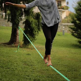 Femme en équilibre sur une slackline verte du kit Spider Slacklines Flyline 25 dans un jardin, démontrant l'utilisation facile et la fiabilité de l'équipement pour la pratique de la slackline, disponible sur NetJuggler