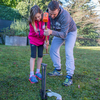 Un homme aide une jeune fille à enfoncer un clou dans le sol à l'aide d'un maillet pour installer un kit d'ancrage de slackline Spider Slacklines dans un jardin. Ils travaillent ensemble pour sécuriser l'installation sur une pelouse verte, entourée de vég