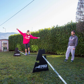 Une jeune fille en sweat-shirt rose marche sur une slackline installée entre deux chavalets Spider Slacklines, sous la surveillance d'un homme. Ils se trouvent dans un jardin spacieux entouré de haies et avec des montagnes visibles à l'horizon. La scène s