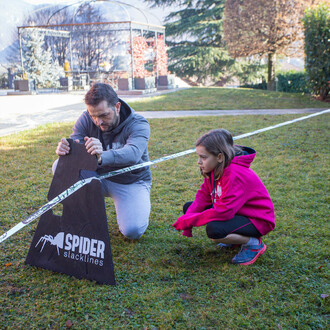 Un homme installe un chavalet pour une slackline Spider Slacklines, observé par une jeune fille en sweat-shirt rose.