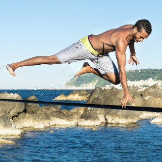 Un homme torse nu en short gris effectue une figure acrobatique sur une slackline tendue au-dessus de l'eau et des rochers en bord de mer. Il est en position horizontale, avec une jambe tendue vers l'arrière et l'autre repliée, une main touchant la slackl