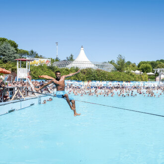 Un homme torse nu en short bleu est assis sur une slackline tendue au-dessus d'une piscine remplie de personnes. Il est en pleine figure acrobatique, les bras étendus pour maintenir l'équilibre. En arrière-plan, une foule de baigneurs profite de la piscin