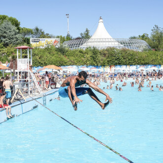 Un homme torse nu en short bleu réalise une figure acrobatique sur une slackline tendue au-dessus d'une piscine bondée. Il est en position accroupie, les mains et les pieds en avant, semblant sauter ou s'accroupir sur la slackline. En arrière-plan, de nom