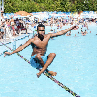 Un homme torse nu en short bleu équilibre sur une slackline tendue au-dessus d'une piscine bondée. Il est en position accroupie, les bras tendus pour maintenir son équilibre. En arrière-plan, de nombreux baigneurs profitent de la piscine et des installati