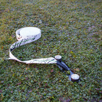 Gros plan sur une sangle de slackline blanche déroulée sur une pelouse, prête à être installée. La sangle est attachée à une ancre au sol, partie du kit d'ancrage pour slackline Spider Slacklines. L'herbe est verte et légèrement humide, indiquant une inst