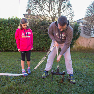 Un homme installe une slackline en fixant la sangle à une ancre au sol , tandis qu'une jeune fille en sweat-shirt rose observe. Ils se trouvent dans un jardin entouré de haies et d'arbres, et la scène est ensoleillée, indiquant une belle journée en extéri