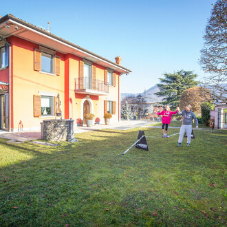 Une jeune fille marche sur une slackline installée dans le jardin d'une maison, avec l'aide d'un homme. Le jardin est spacieux, entouré de haies et d'arbres, et la maison en arrière-plan est de couleur orange avec des volets marron. La scène se déroule pa