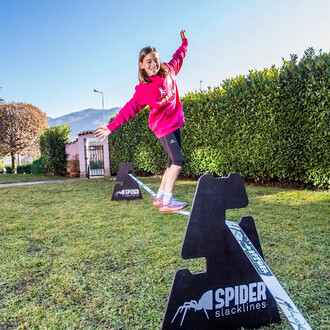 Une jeune fille en sweat-shirt rose marche sur une slackline installée entre deux chavalets Spider Slacklines dans un jardin. Elle sourit et tend les bras pour maintenir l'équilibre. Le jardin est entouré de haies et la scène se déroule par une journée en