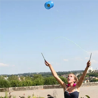 Une personne utilise un diabolo bleu en plein air, levant les bras avec des baguettes en superglass. L'arrière-plan présente un paysage verdoyant sous un ciel dégagé.
