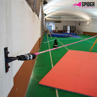 Photo d'une sangle de slackline Spider Slacklines rose installée dans une salle de sport. La sangle est attachée à un mur avec un support métallique noir et passe au-dessus d'un sol en mousse de protection verte et orange. Le logo Spider Slacklines est vi