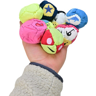 Une main tenant un assortiment de neuf boules colorées en tissu, aux motifs variés, y compris des visages souriants et des étoiles, représentant les TSHAKA du kit pédagogique. La main est partiellement couverte par une manche de vêtement en fourrure.