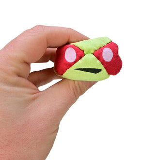 Un utilisateur tient un TSHAKA en forme de petit personnage, avec un corps vert et des yeux roses. Le TSHAKA est manipulé à une main, mettant en avant sa taille compacte et sa texture douce.