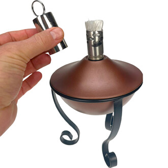 Main retirant le bouchon métallique près du brûleur d’une lampe à huile torchière en métal brun, avec mèche visible et support en ferronnerie décorative.