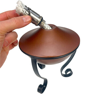 Plan rapproché montrant une main insérant une mèche tressée dans le brûleur métallique d’une lampe à huile torchière, avec le corps brun et le support en métal sombre.