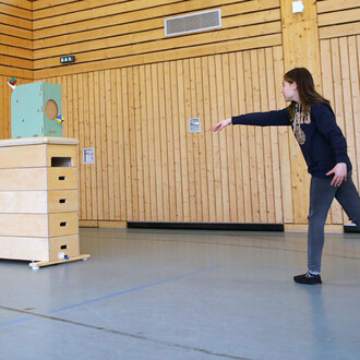 Image d'un enfant lançant une balle vers une machine de lance-balles Pedalo® dans une salle de sport. La machine est en bois, a un trou sur le devant et des éléments colorés sur les côtés. Des murs en bois complètent l'arrière-plan.