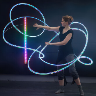 Femme en train de jongler avec un Levistick lumineux. Le bâton est illuminé de couleurs vives et crée des motifs lumineux dans l'air. La personne porte un haut noir et un pantalon gris, avec des chaussettes assorties. L'arrière-plan est sombre, mettant en