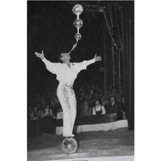 Un artiste de cirque en costume brillant équilibre sur une boule à facettes tout en faisant des bulles avec de l'eau, sous un éclairage de scène. Le public est visible en arrière-plan, admirant la performance.