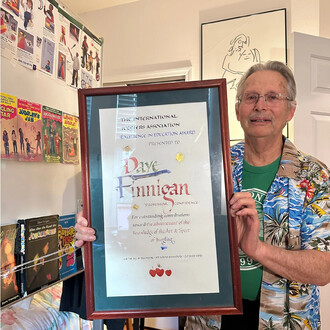 Homme âgé portant une chemise hawaïenne tient un cadre avec un diplôme encadré, dans une pièce décorée de posters de jonglage et de livres sur le jonglage sur une étagère.