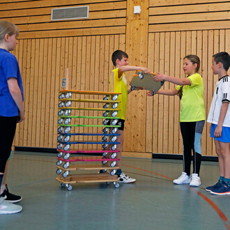 Deux enfants en t-shirts jaunes échangent un plateau sur roulettes dans une salle de sport. Un enfant en t-shirt bleu observe, tandis qu'un autre enfant se tient à droite, portant un maillot à rayures. En arrière-plan, un support coloré est visible, compo