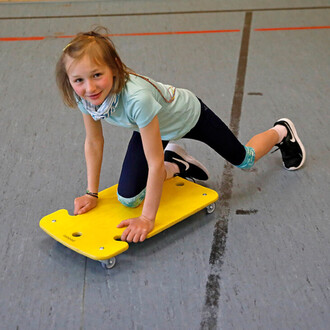 Une fille en train de jouer sur un skateboard jaune à roulettes, sur un sol de gymnase. Elle est à quatre pattes, souriante et porte un t-shirt bleu clair, un pantalon noir et des chaussures de sport noires.