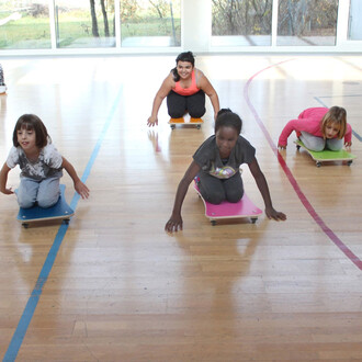 Quatre enfants s'amusent sur des planches à roulettes colorées dans une salle de sport. Ils sont inclinés en avant, les bras tendus, en train de glisser sur le sol en bois. Au fond, de grandes fenêtres laissent entrer la lumière naturelle. Les enfants por