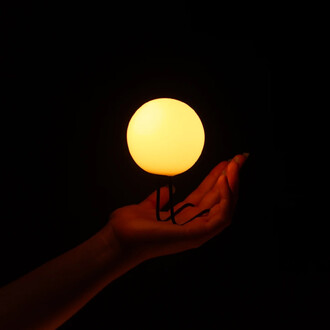 Une main tenant une boule lumineuse orange, placée sur un support, le tout sur fond noir.