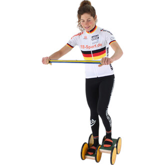 Femme souriante portant un maillot de cyclisme avec des couleurs allemandes, en train de jongler sur un matériel de jonglerie avec des roulettes tout en tenant une bande de résistance.