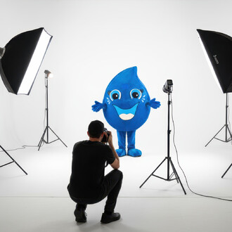 Mascotte Goutte d’eau bleue en costume, souriante et aux bras ouverts, photographiée en studio avec un fond blanc et des lumières de photographie.
