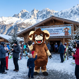 Mascotte mouflon malicieux en costume brun avec grandes cornes crème, saluant les enfants lors d’une fête hivernale alpine devant un chalet en bois et un panneau Festival d’Hiver des Alpes, entourée de spectateurs et de skieurs.